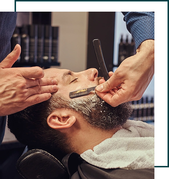 Bearded male sitting in an armchair in a barber shop while hairdresser shaves his beard