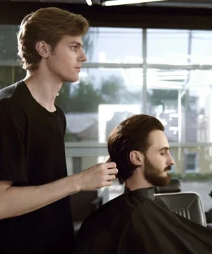 Barber works in barbershop. Handsome man with dark hair, beard and moustache has haircut in hairdressing saloon.